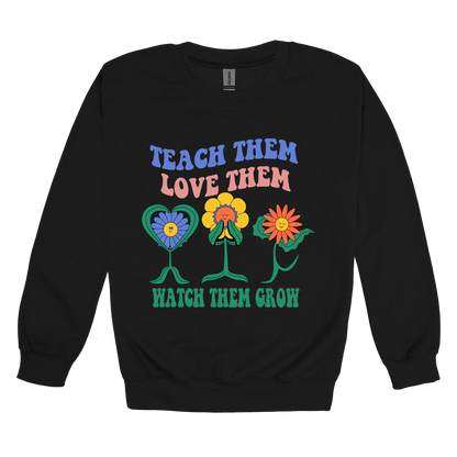 Teach Them Love Them teacher design on Black adult Crewneck, front view.
